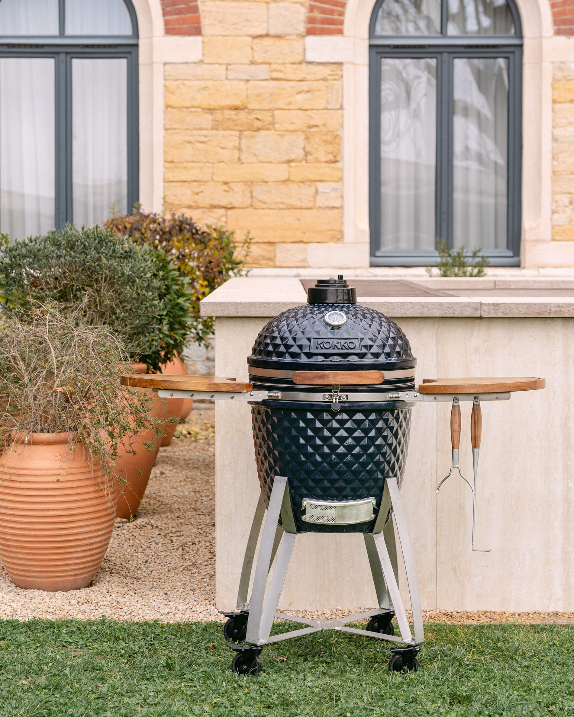 Vue d’ensemble du kamado Kokko sur chariot mobile avec plateaux rabattables et ustensiles suspendus, en situation extérieure.