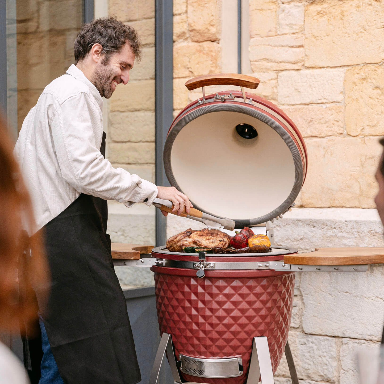 Un utilisateur cuisine de la viande et des légumes sur un kamado Kokko rouge en céramique, couvercle ouvert, lors d’un moment convivial en extérieur.