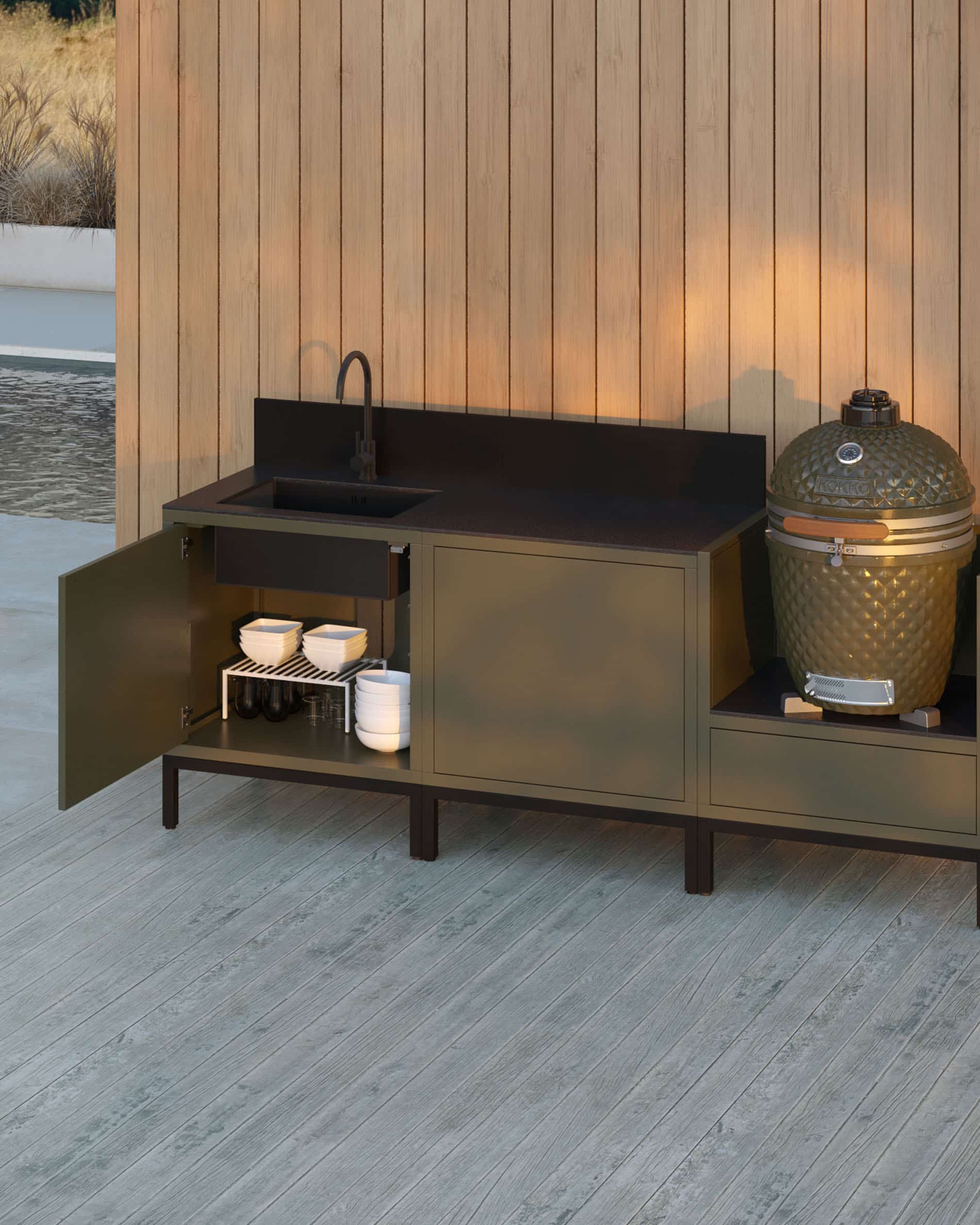 Cuisine pilotis 3 modules pour kamado avec évier intégré, meuble coloris olive avec plan de travail et crédence noirs, porte du module gauche ouverte révélant un rangement intérieur, évier avec robinet à gauche, deux modules fermés au centre et kamado intégré dans le module latéral droit, structure sur pieds noirs, installation en extérieur contre un mur en bardage bois.