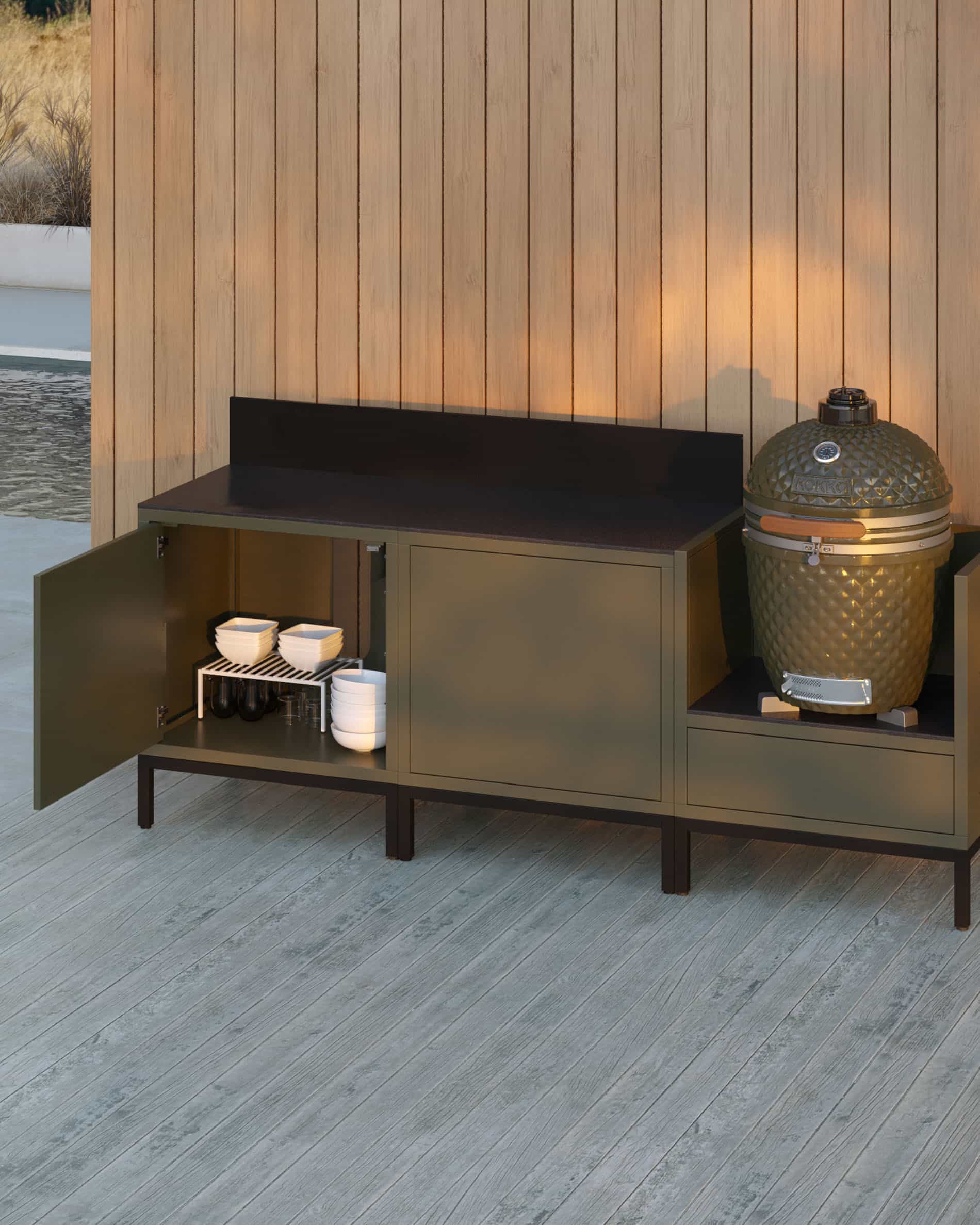 Outdoor-Küche auf Stelzen mit 3 Modulen für Kamado, Möbel in Olivgrün mit dunkler Arbeitsplatte und passender Rückwand, geöffnete Aufbewahrungstür zeigt gelagertes Geschirr, Kamado im rechten Seitenmodul integriert, Konstruktion auf schwarzen Füßen.