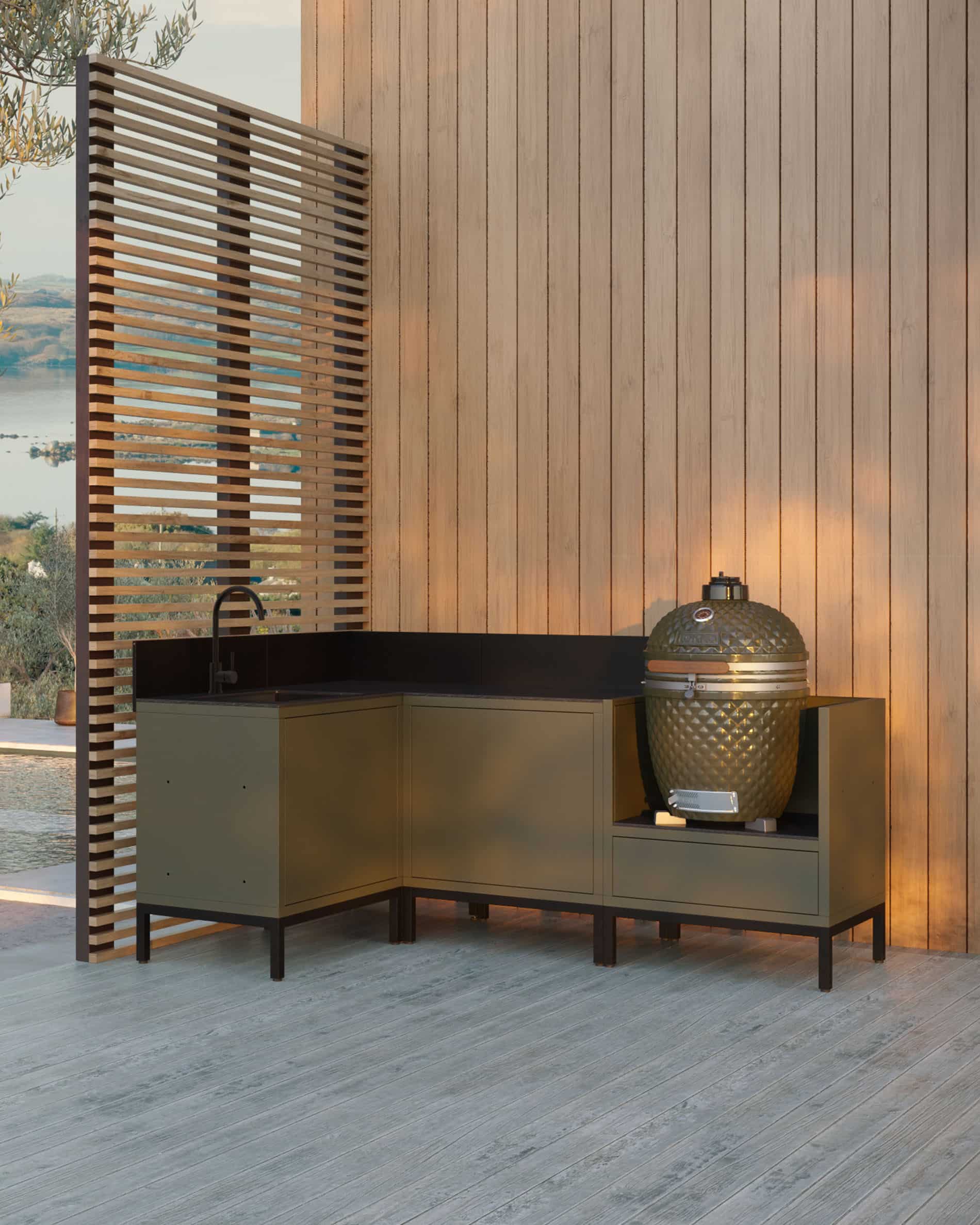 Cuisine pilotis d’angle coloris olive composée de quatre modules avec évier intégré, plan de travail noir et emplacement dédié au kamado, installée sur une terrasse en bois contre un bardage vertical.