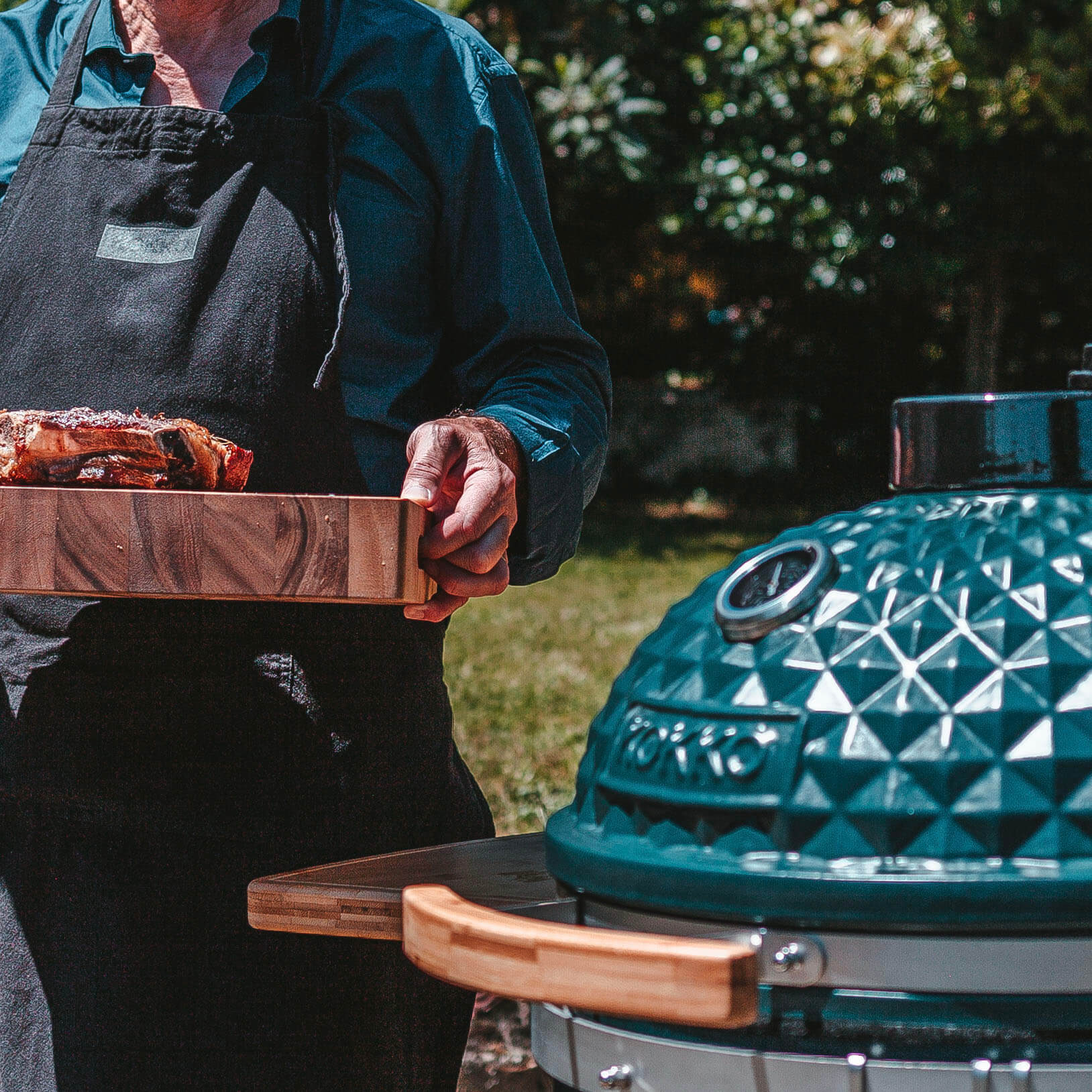 Kamado Kokko en céramique, fermé, avec tablettes en bois, posé en extérieur, à côté d’une personne tenant une planche en bois avec une pièce de viande grillée.