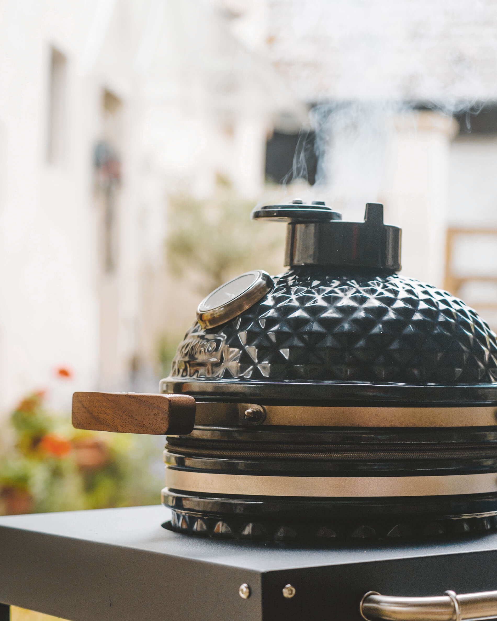 Kamado Kokko coloris noir posé sur chariot, avec couvercle en céramique texturée, poignée en bois, thermomètre intégré et fumée s’échappant de la cheminée.
