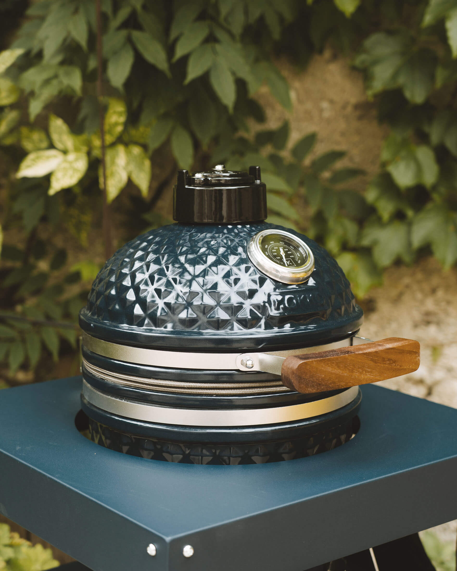 Vue de face d’un kamado Kokko coloris océan posé sur chariot, mettant en valeur la céramique texturée, le thermomètre intégré et la poignée en bois.