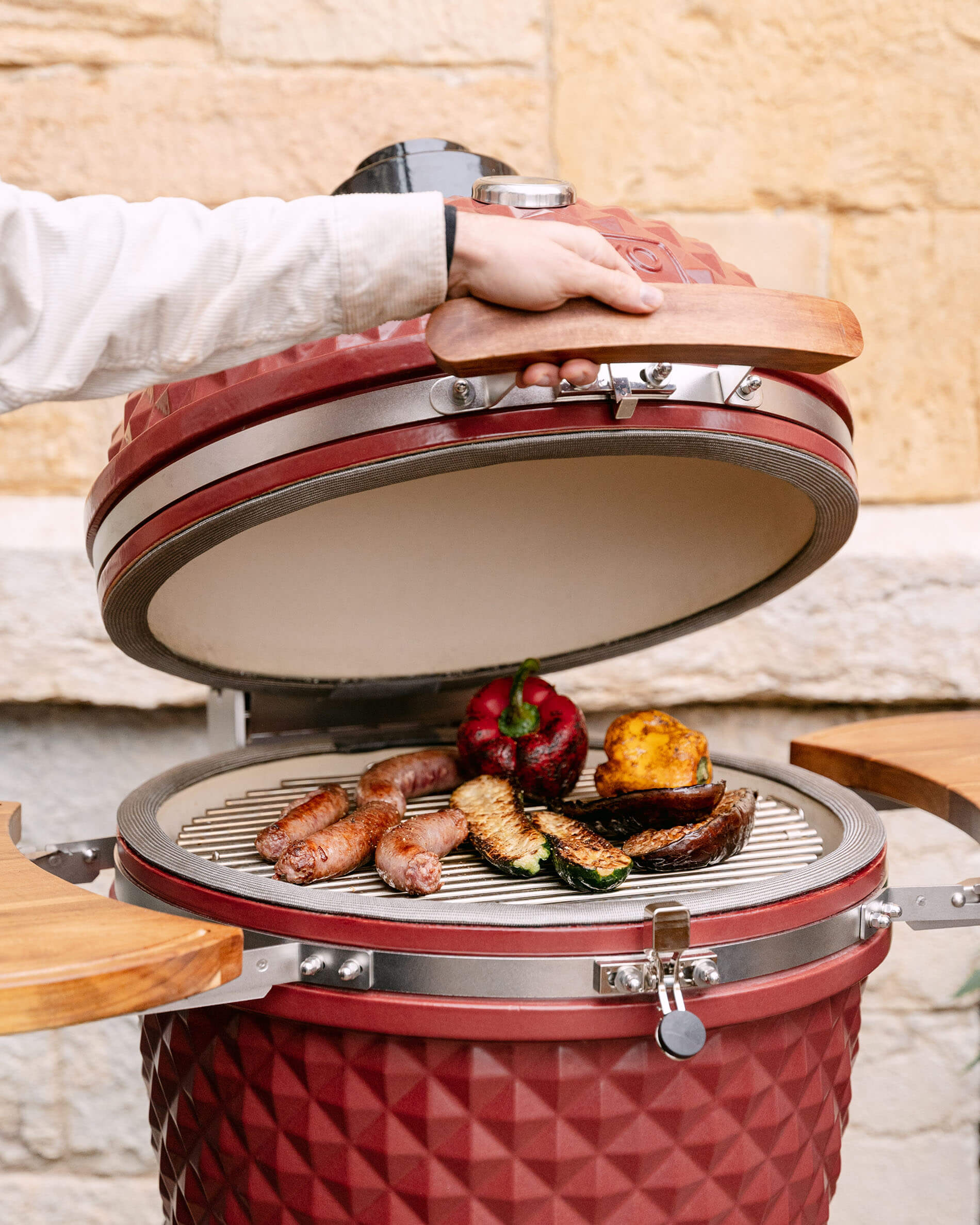 Kamado Kokko 22 pouces en céramique rouge oxyde ouvert, avec cuisson de saucisses et légumes sur la grille.