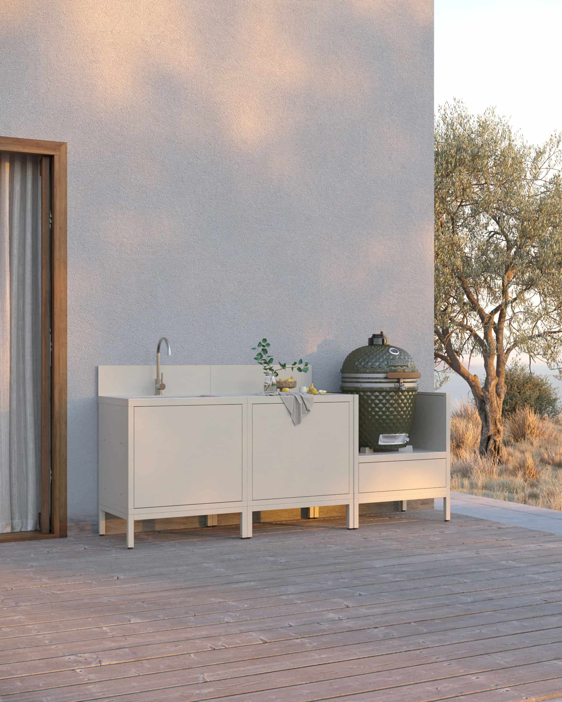 Cuisine pilotis coloris crème composée de trois modules avec évier intégré, plan de travail clair et emplacement dédié au kamado, installée sur une terrasse en bois.